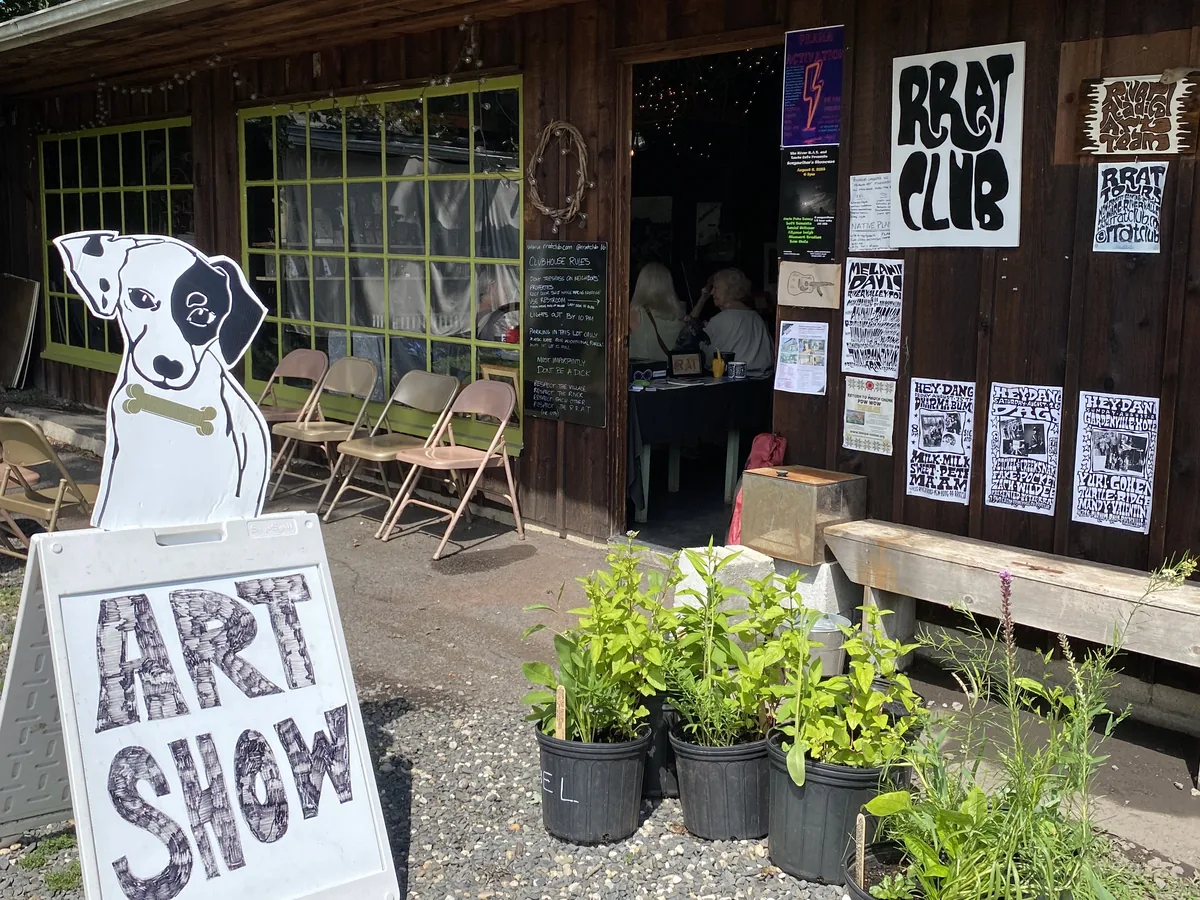 A hand-lettered ART SHOW sandwich board, topped with a cut-out of a black-and-white dog holding a bone, stands on gravel beside black pots of leafy plant starts outside a dark wood-sided building hung with posters.