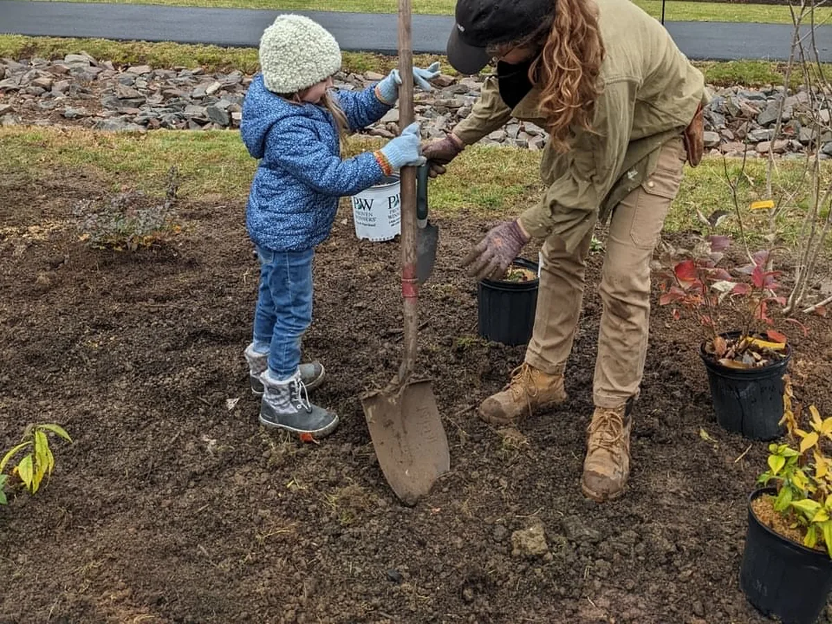 A small child in a blue puffy jacket and cream knit hat holds a young tree upright while an adult in a tan work jacket steadies it and reaches for a nursery pot, on a bed of freshly turned soil.